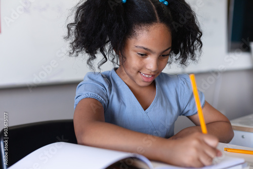 Smiling biracial elementary schoolgirl writing on book while studying at desk in classroom