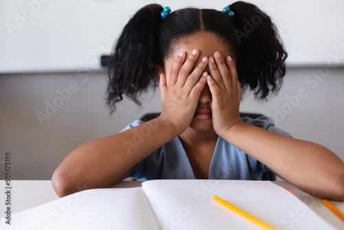Depressed biracial elementary schoolgirl with hands covering face sitting at desk in classroom