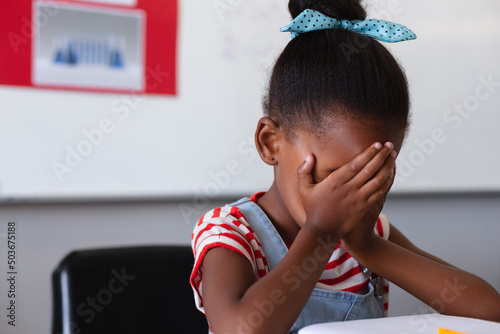 Sad african american elementary schoolgirl hands covering face sitting at desk in class