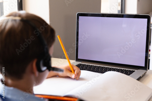 Caucasian elementary schoolboy writing on book while using laptop with copy space at desk in class