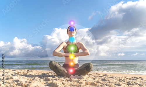 Slim woman meditating in lotus position in nature. Seven glowing chakra symbols representing spiritual health, harmony, and balance against a blue sky.