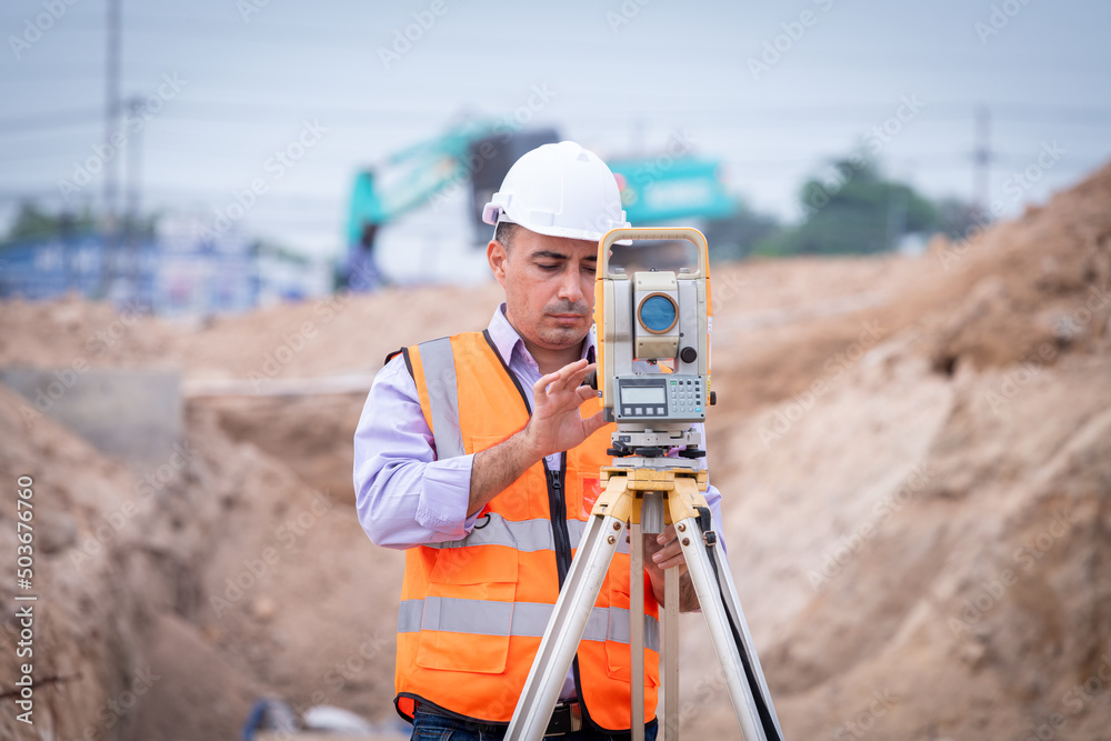 Surveyor engineer wearing safety uniform ,helmet and radio communication with equipment theodolite to measurement positioning on the construction site of the road with construct machinery background Surveyor engineer wearing safety uniform ,helmet and radio communication with equipment theodolite to measurement positioning on the construction site of the road with construct machinery background