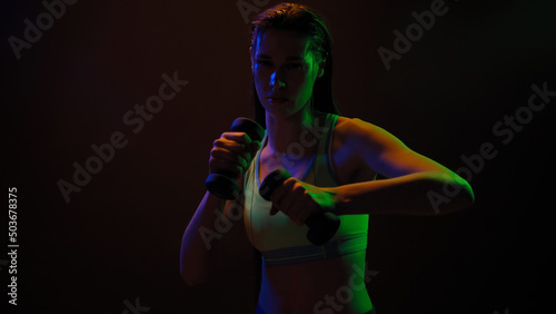 Sportive girl posing doing hand workout biceps with dumbbells standing in neon lights wearing sport top working on a arm weight hit isolated on black background. Neon light concept