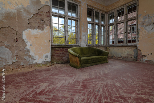 Obraz na plátně Old dusty green sofa in a ruined room with brick destroyed walls, scratched floors and windows without glass