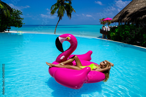 Beautiful sexy tanned woman on pink flamingo in pool. Young glamour girl in swimsuit on Maldives island. Perfect body bikini model in luxury resort on Maldives. Luxury travel. Summertime. © marcink3333