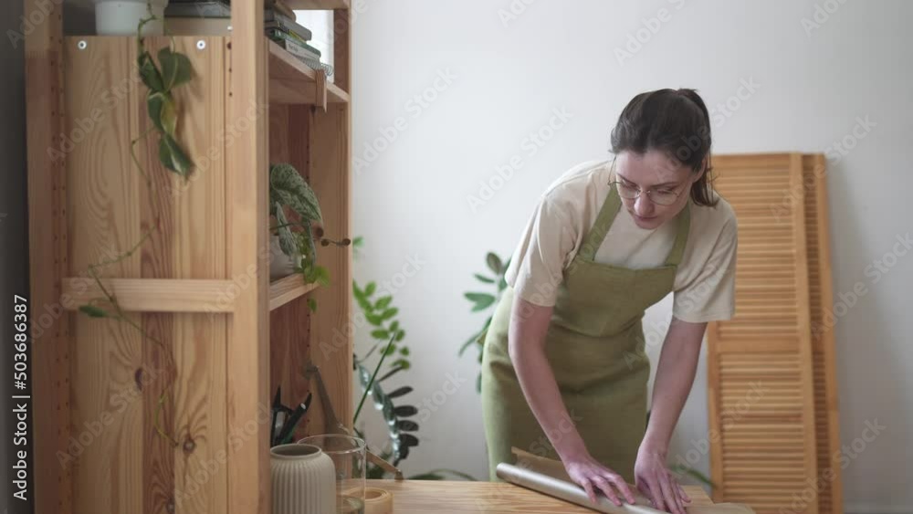 A florist in apron unwraps a craft wrapping paper. Flower and gift shop. Female Seller A florist in apron unwraps a craft wrapping paper. Flower and gift shop. Female Seller