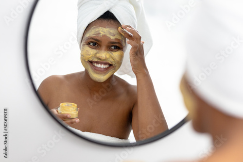 Smiling african american mid adult woman looking in mirror and applying face pack photo