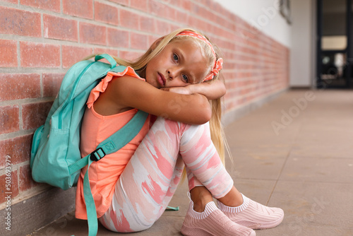 Portrait of caucasian elementary schoolgirl with backpack sitting on floor in school corridor