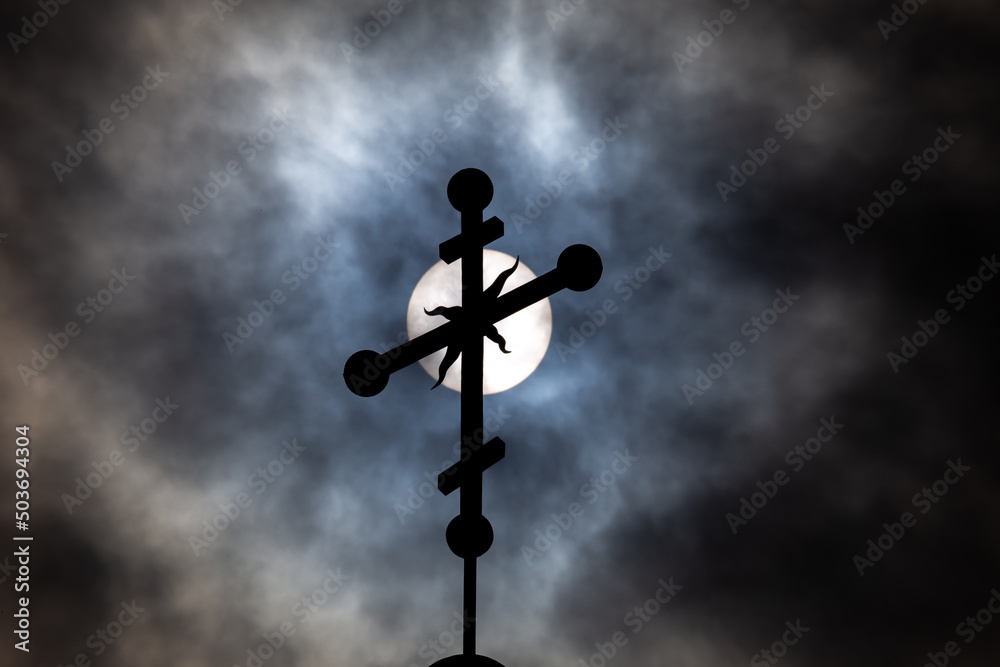 Orthodox cross on top of the church against the dramatic sky. Photo taken against the sun behind the clouds.