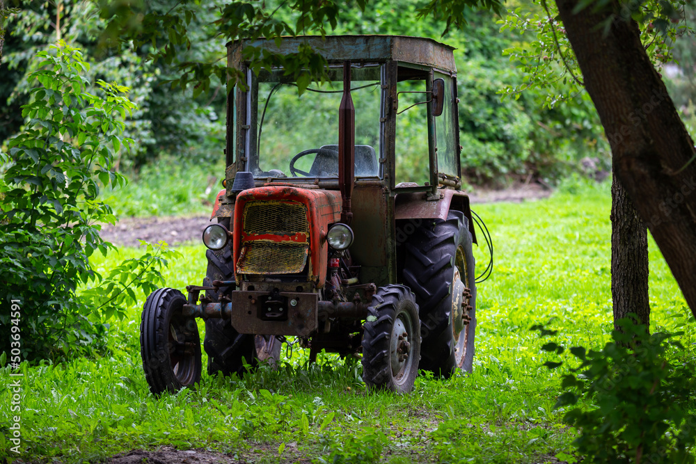 An old red tractor standing in the shade among the trees. The photo was taken on a cloudy day with good lighting conditions. An old red tractor standing in the shade among the trees. The photo was taken on a cloudy day with good lighting conditions.