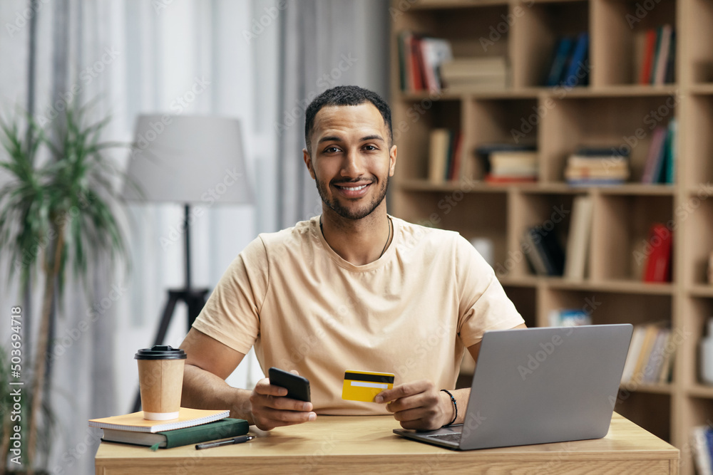 Obraz premium Cheerful arab man purchasing goods on web using smartphone, laptop and credit card, enjoying shopping in internet