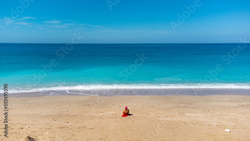 Aerial view of woman in red dress, sitting on the sandy beach, enjoying soft turquoise ocean wave. Tropical sea in summer season on Megali Petra beach on Lefkada island.