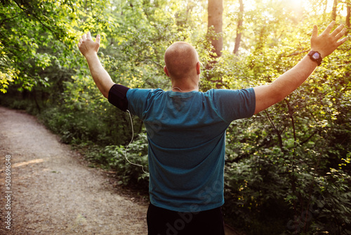 Sporty man raising arms towards beautiful sunset in the public park. Young man wearing blue shirt and black shorts runs while enjoying freedom on an idyllic road during sunset surrounded by nature.