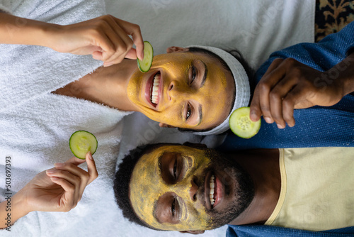 Happy multiracial young couple with facial mask holding cucumber slices while relaxing on bed photo