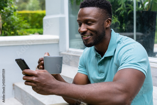 Smiling african american young man using smart phone while having coffee on steps outside house