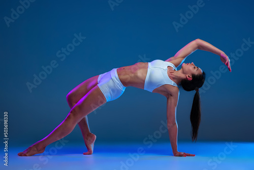 Portrait of young soprtive woman training isolated over blue studio background in neon light. Stretching exercises