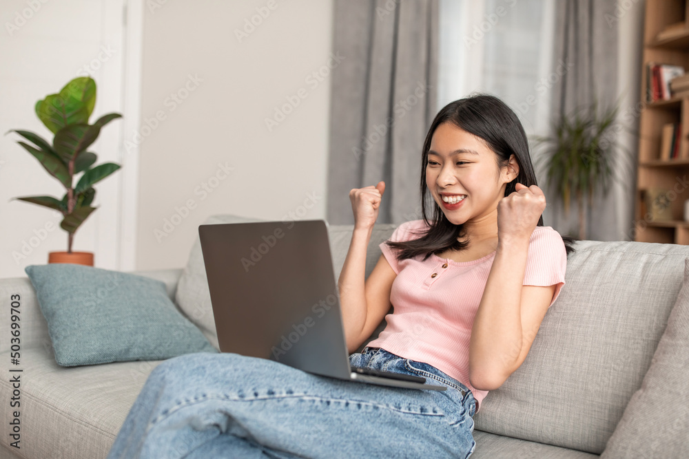 Excited korean lady sitting on couch with laptop, lifting hands up, gesturing YES and celebrating success at home Excited korean lady sitting on couch with laptop, lifting hands up, gesturing YES and celebrating success at home