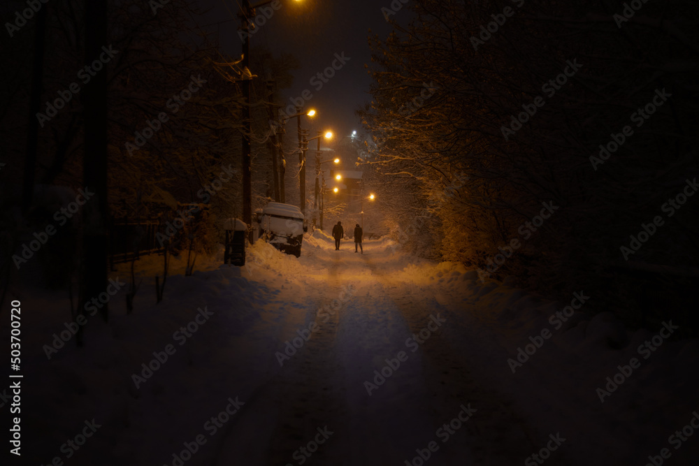 Evening winter road, two people in the distance. Snowfall during winter. Beautiful winter street evening landscape. Beautiful winter night. 