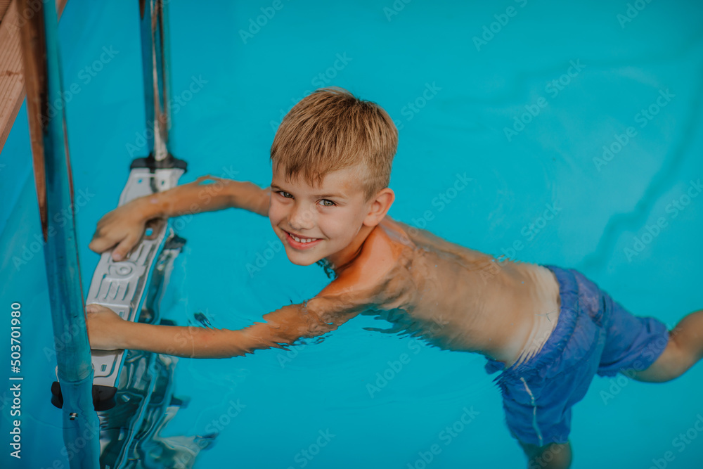 Cute young boy holds pool's stairs and looking into the camera. Copy space. Summertime concept.