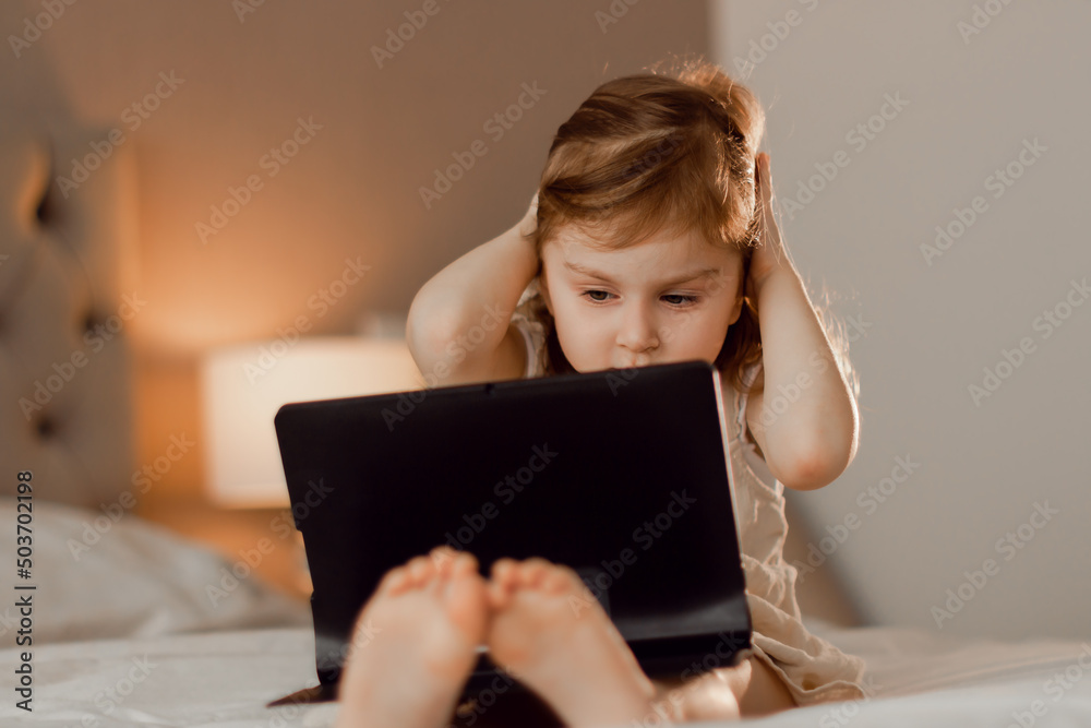 Cute toddler little girl wearing casual dress sitting lying in bedroom with modern interior in beige colors using laptop tablet pad. Cute toddler little girl wearing casual dress sitting lying in bedroom with modern interior in beige colors using laptop tablet pad.