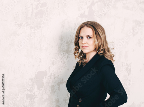 A beautiful young woman of Caucasian appearance with blond hair, in a business suit, looks at the camera, smiles. Business lady. good-looking beautiful woman isolated against blank gray wall.