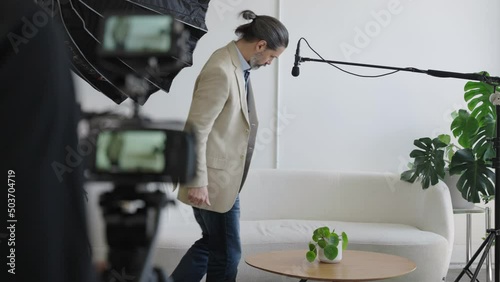 Male Business Man Walks Into Video Production Set and Sits Down Ready to be Interviewed. Behind the Scenes Camera, Lighting and Audio Equipment Can Be Seen. Slow Motion Scene of On Camera Talent