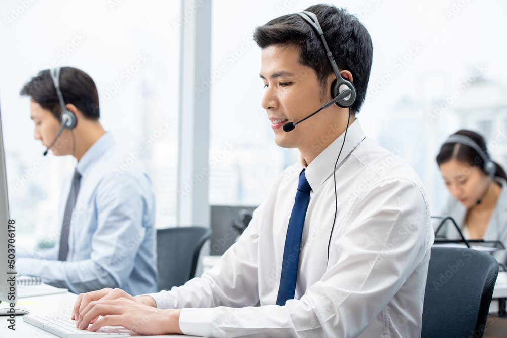 Side view of focused Asian male worker in headset typing on computer while working in call center with coworkers Side view of focused Asian male worker in headset typing on computer while working in call center with coworkers