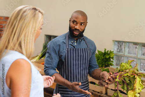 African american mid adult bald vendor showing beets to caucasian female customer in store