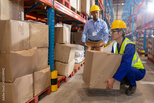 African american young foreman guiding asian mature male worker while lifting cardboard box by shelf photo