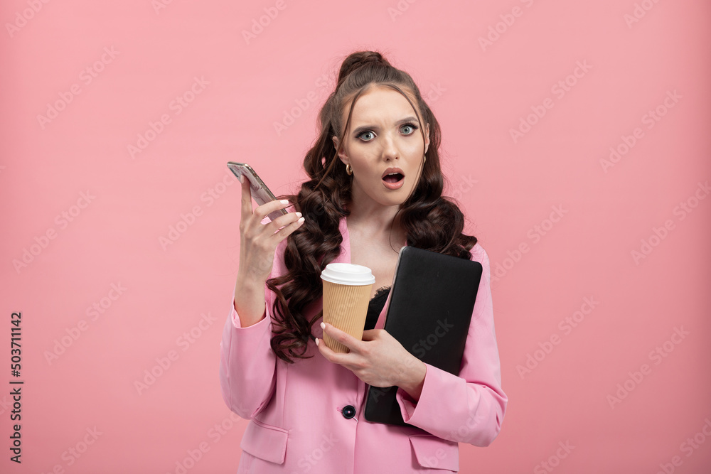 Portrait of shocked, puzzled girl in jacket reading post on social networking site using phone, chatting, holding laptop and coffee mug looking puzzled. Studio shot isolated on pink background. Portrait of shocked, puzzled girl in jacket reading post on social networking site using phone, chatting, holding laptop and coffee mug looking puzzled. Studio shot isolated on pink background.