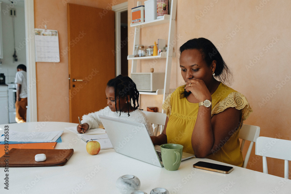 © Maskot - Female freelancer with daughter working on laptop at home
