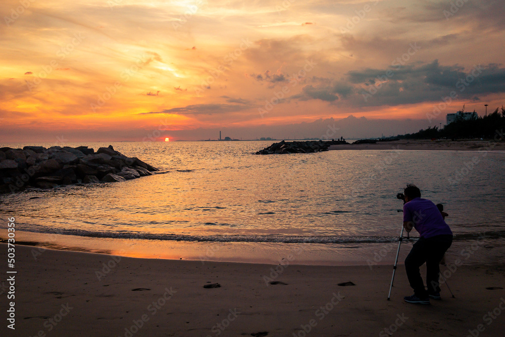 A man standing on the beach behind the camera on the tripod. The camera point to cloudy sky and sunset by the sea. A man standing on the beach behind the camera on the tripod. The camera point to cloudy sky and sunset by the sea.