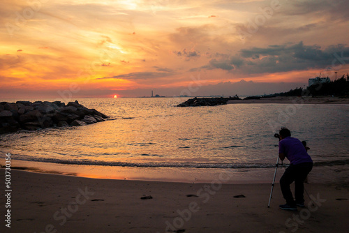 A man standing on the beach behind the camera on the tripod. The camera point to cloudy sky and sunset by the sea.
