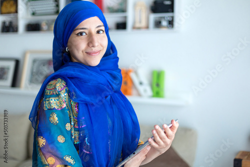 Smiling young woman at home wearing typical traditional Muslim dress. She is holding a digital tablet. Space for text.