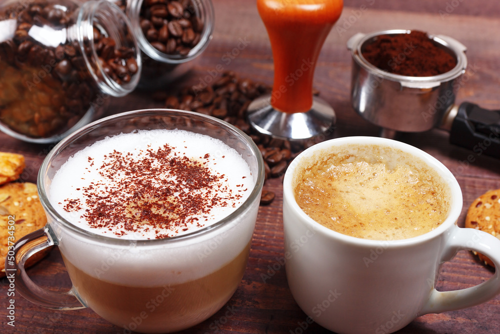 Cups of espresso and cappuccino, crackers, holder with ground coffee, tamper and coffee beans on wooden table. Cups of espresso and cappuccino, crackers, holder with ground coffee, tamper and coffee beans on wooden table.