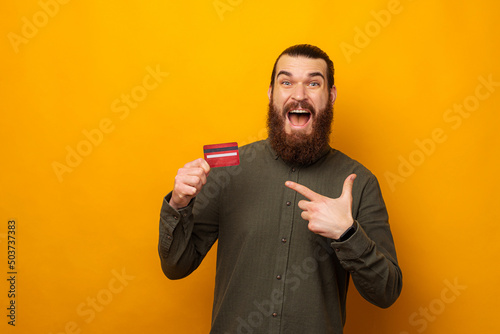 Shocked young bearded man is pointing at the credit card he is holding.