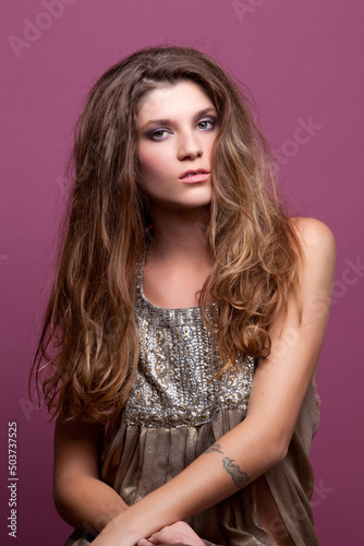 Beautiful Young Glam Punk Rock Brunette Fashion Model Posing in Metallic Clothing with Tattoos in Studio against Pink Background Looking at Camera