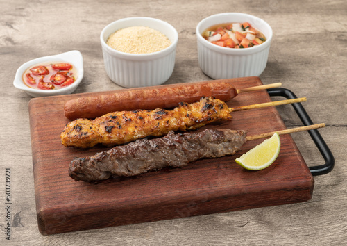 Meat, chicken and pork sausage skewers over wooden board with farofa and vinaigrette