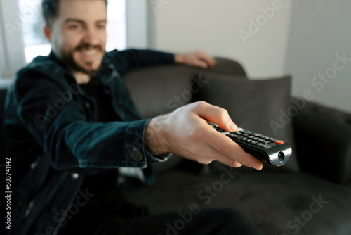 Happy young man wearing denim clothes with remote control switching channels on TV at home. Happy young guy watching interesting program on television indoors.