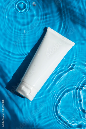 Plastic white tube for cream or lotion on beautiful burst and glare. Skincare or sunscreen cosmetic on blue background. Beauty concept for face care. Natural sunlight and water spills. Summer mood.
