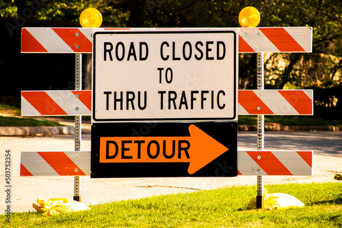 Road Closed to thru traffic Detour sign on striped barrier with reflectors on side of road on bright sunny day
