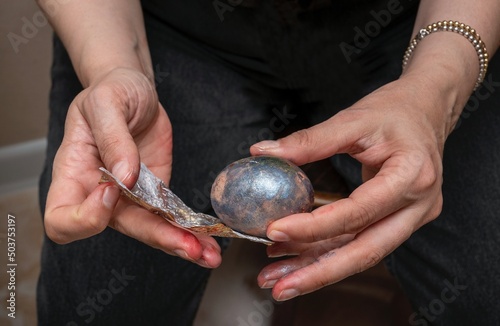 Close-up of white-skinned female hands holding foil and a dark-colored chicken egg in the process of making Easter decorations for the holiday