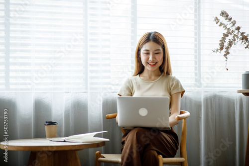 Young Asian lady studying online from home, reading a book and using laptop pc, sitting on the coffee shop, free space. E-learning, web-based education concept