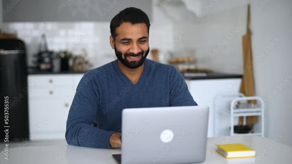 Young indian male employee is using video connection for virtual meeting with co-workers, eastern guy working remotely from home,sits in front of laptop in the kitchen, greeting and waving into webcam