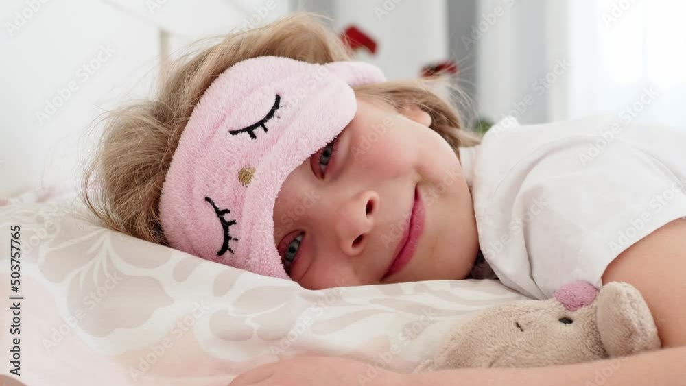 girl sleeping. A beautiful, sweet little girl with long hair is lying in bed hugging a soft toy rabbit. The eyes are covered with a sleep mask. The child woke up, took off his mask and smiled a happy 