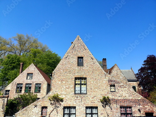 Blick von der Brücke auf den Beginenhof mit alter Mauer aus Naturstein und altem Backsteinbau mit Spitzgiebel vor blauem Himmel im Sonnenschein in der Altstadt von Brügge in Westflandern in Belgien