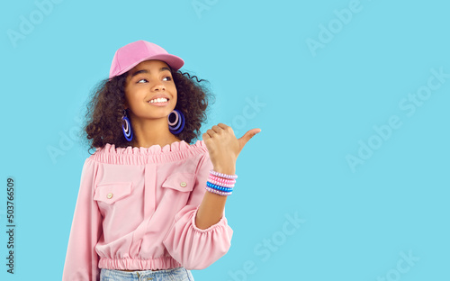 Papier peint Black girl with curly hair wearing pink baseball hat, blouse, bracelet and earrings standing and pointing sideways at copy space on blue background