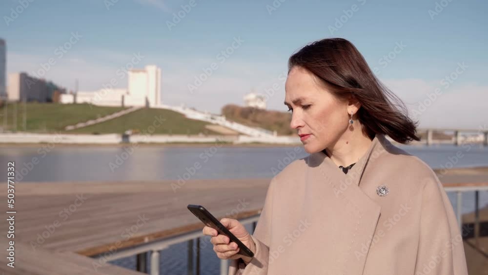 A business lady in a coat looks through messages on a mobile phone on the street. Girl in a coat on the shore of a reservoir in the city with a smartphone in her hand. A business lady in a coat looks through messages on a mobile phone on the street. Girl in a coat on the shore of a reservoir in the city with a smartphone in her hand.