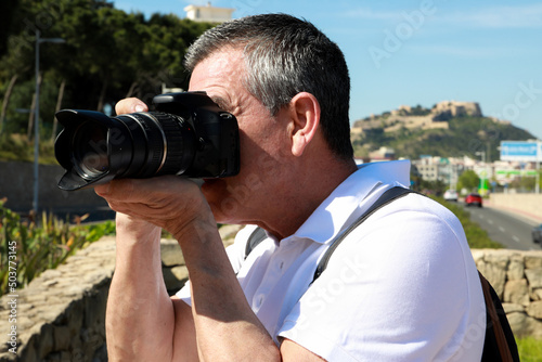 A 60 year old man wearing sunglasses, on summer vacation sightseeing in the city of Alicante, taking pictures with a camera in front of the Santa Barbara Castle.