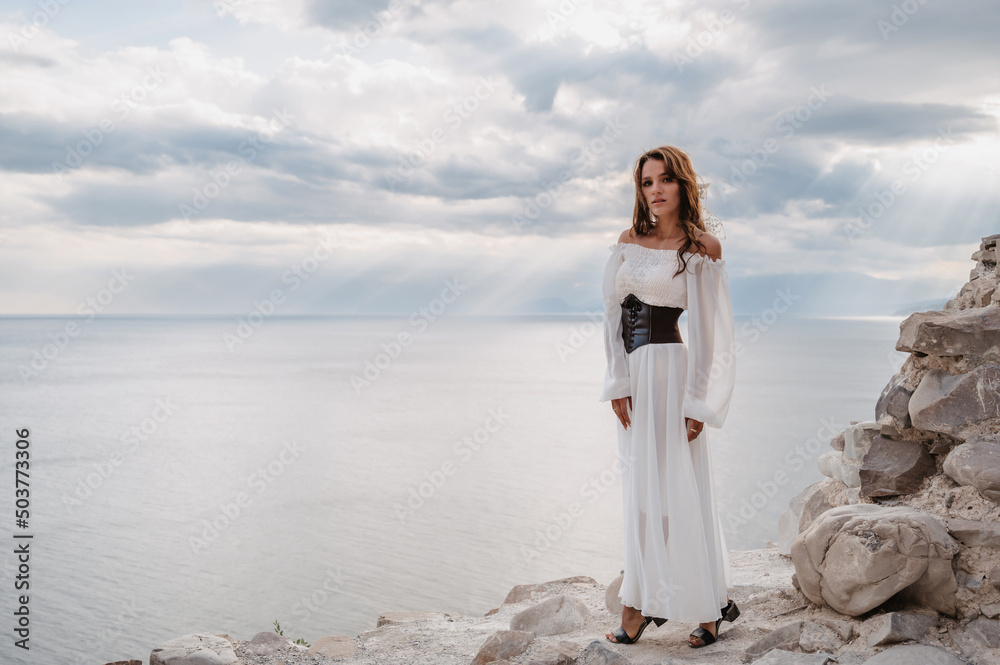 A young woman in a white dress stands barefoot on a cliff face in full height. The dress flutters in the wind. A sacred glow breaks through the clouds. The girl looks like an angel